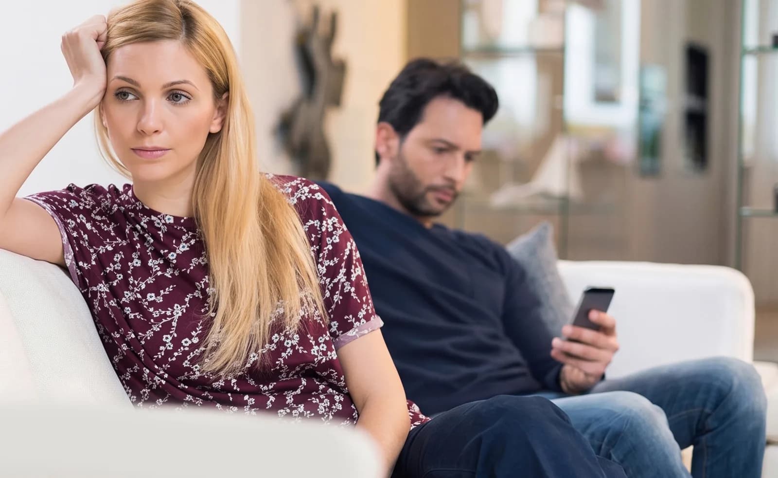Casal sentado no sofá; o homem olha para o celular enquanto a mulher demonstra distanciamento emocional.